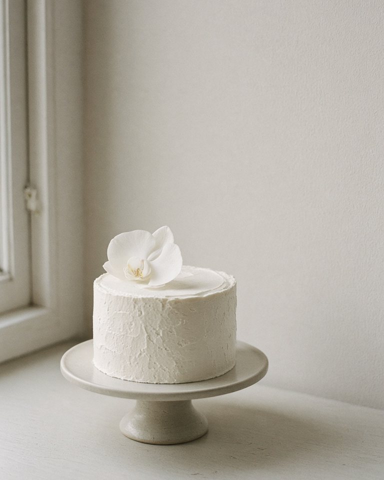 A simple white cake with one orchid on a stand by a window